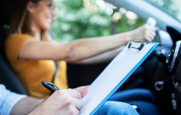 close-up-view-driving-instructor-holding-checklist-while-background-female-student-steering-driving-car